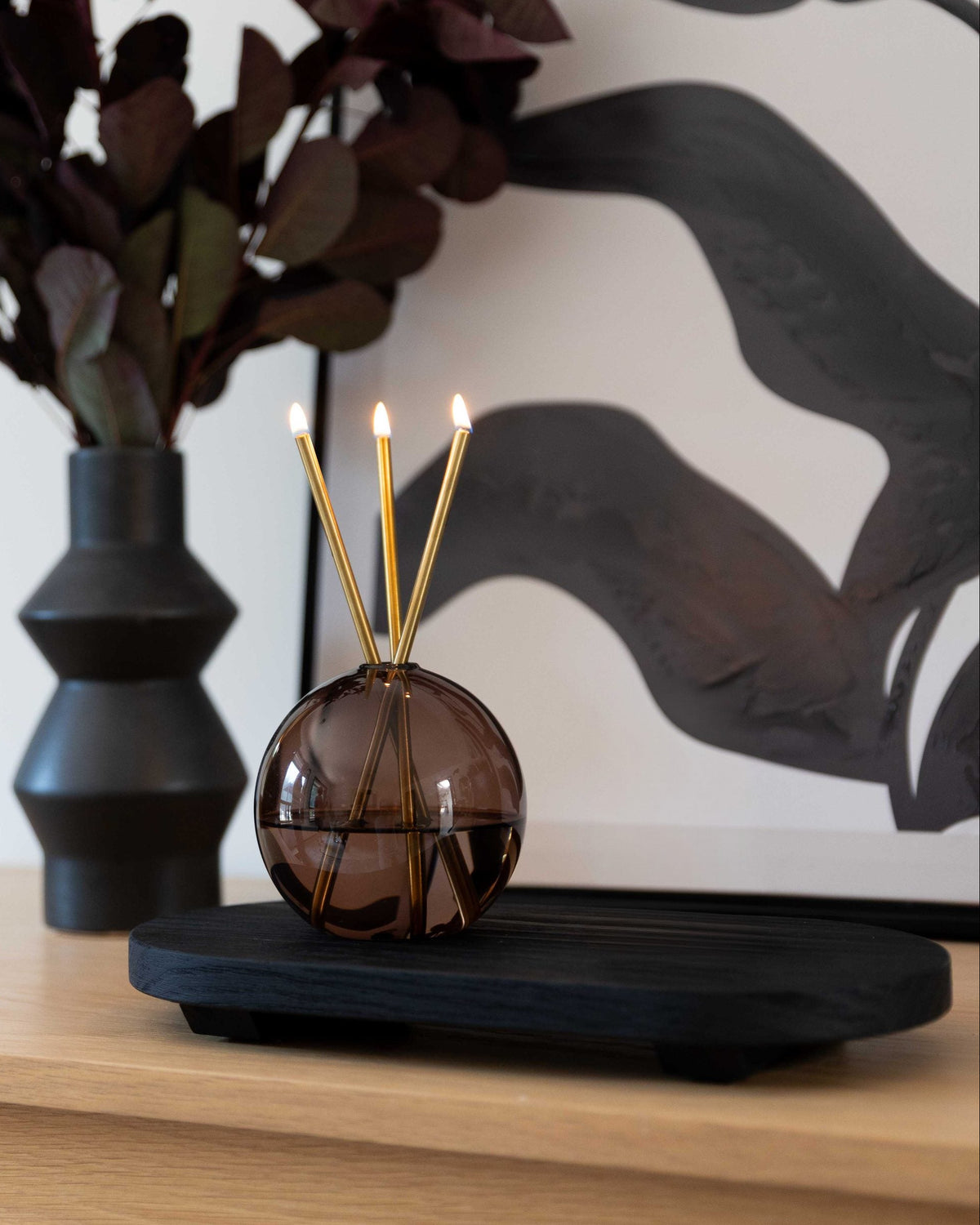 A spherical amber glass reed diffuser with three lit gold reeds sits on a black wooden tray on a light wood surface, with a dark vase and abstract wall art in the background.