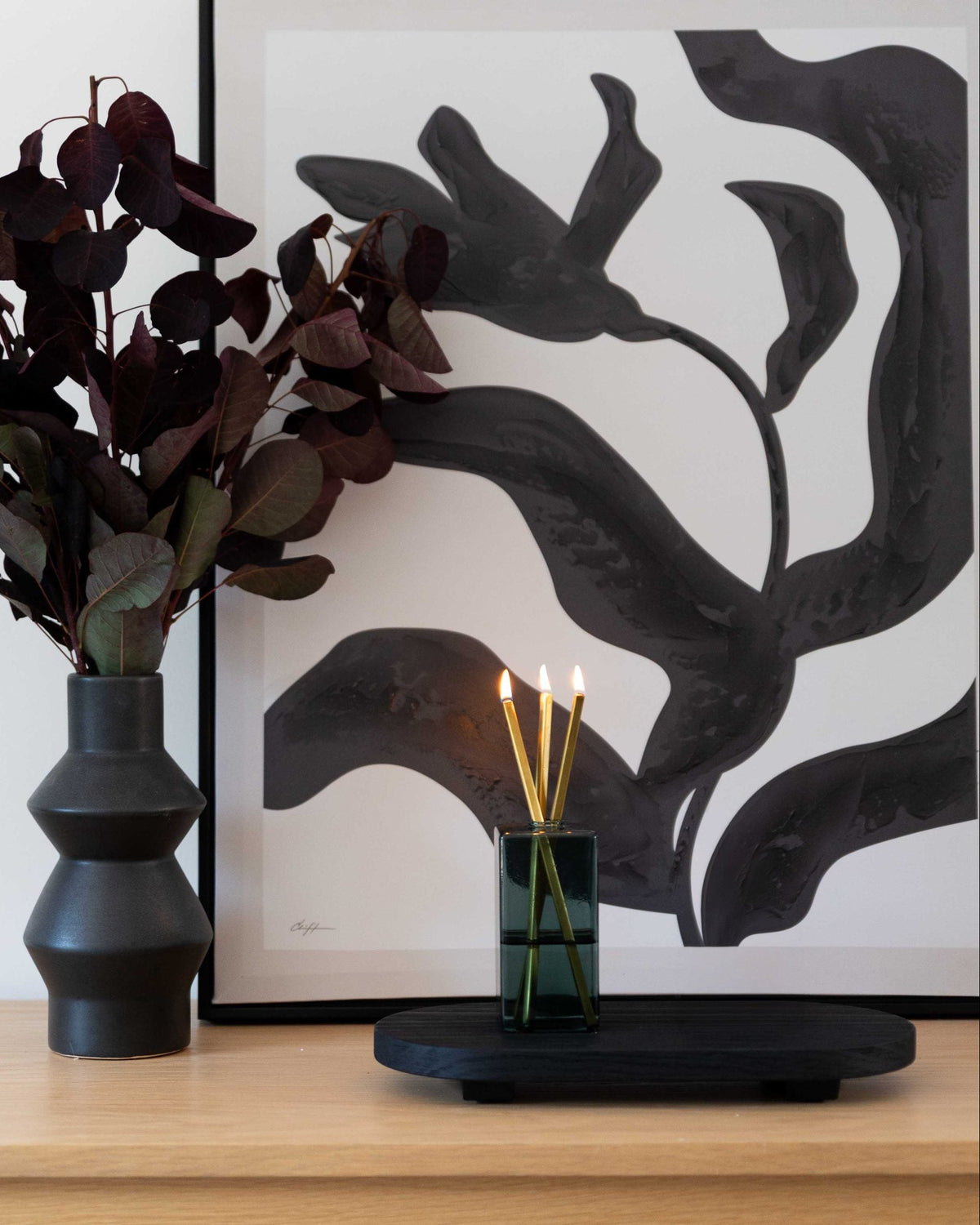 A styled interior scene showing a dark charcoal ceramic vase with dark burgundy dried leaves, next to a black tray holding a green glass Everlasting Candle Set with three lit Everlasting Candles, all set against a large black and white abstract art print on a light wood wall.